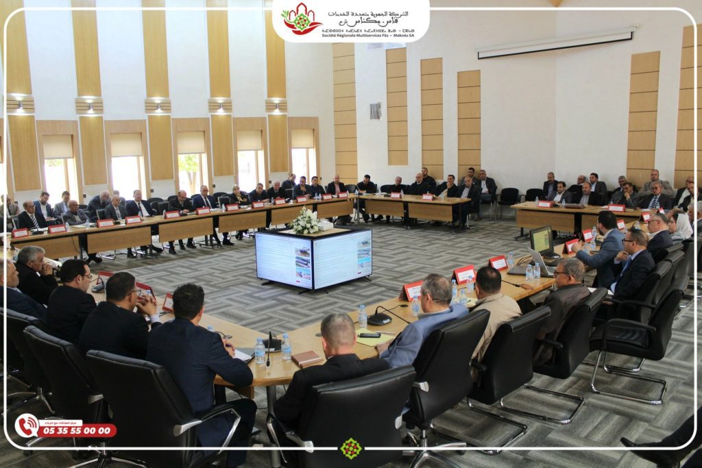 1775660614191 | MCG24 | موقع إلكتروني مغربي شامل متجدد على مدار الساعة Large conference room with many attendees in suits seated around a U-shaped table, a central screen showing slides, and a decorative banner/logo at the front.