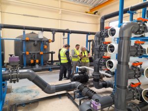1776682567634 | MCG24 | موقع إلكتروني مغربي شامل متجدد على مدار الساعة Three workers in high-visibility vests discuss piping equipment in a large industrial pump room with large grey pipes and a spherical tank in the background.