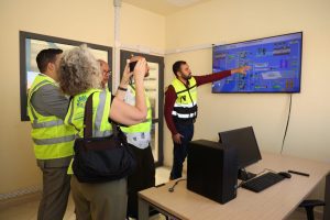 1776682567706 | MCG24 | موقع إلكتروني مغربي شامل متجدد على مدار الساعة Group of people in high-visibility vests gather around a wall-mounted monitor, with one person pointing at a complex process diagram on screen in a control room.