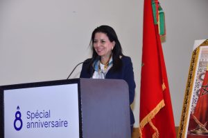 Woman speaks at a podium during an anniversary event; a red flag and a framed artwork are visible beside her, podium sign reads '8e Spécial anniversaire'