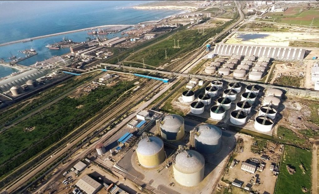 4865 | MCG24 | موقع إلكتروني مغربي شامل متجدد على مدار الساعة Aerial view of an industrial complex with numerous large cylindrical storage tanks near the coast, connected by rail lines and roads across a grassy landscape.