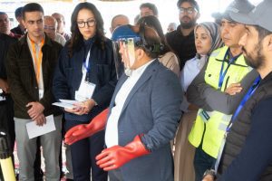 Group of professionals observing a safety demonstration; a man in a suit wears a clear face shield and oversized red gloves as he speaks.