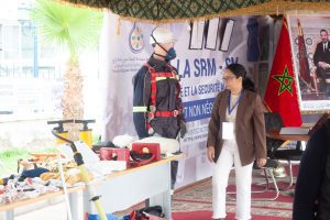 Mannequin in safety harness and helmet beside a display of PPE at a safety expo with a woman in a blazer nearby.