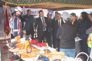 Speaker in a suit addresses a group under a decorated tent, with a firefighter mannequin and safety gear on display nearby.