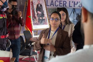 Woman in brown blazer and glasses speaks to an audience, gesturing with hands; camera crew and other attendees nearby, Moroccan flag and a framed portrait in the background.