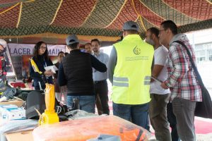 Group of people under a colorful canopy discussing safety gear at a booth, with equipment on the table nearby.