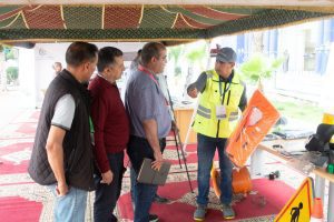 Instructor in a yellow safety vest demonstrates an orange life jacket to a group of men inside a tent, on a red carpet