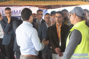Group of professionally dressed men in a discussion under a striped tent; a man in a bright yellow safety vest is on the right side of the frame.