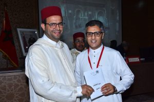 Two men in traditional Moroccan dress (white robes and red fez) smile as one presents a certificate to the other at a formal ceremony.