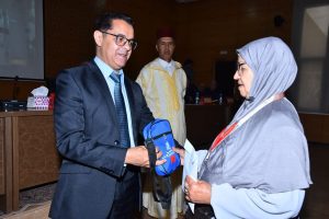 Man in a dark suit hands a small blue pouch to an elderly woman wearing a hijab at a formal ceremony.”
