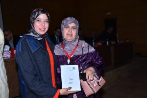 Two women in headscarves pose for a photo at a conference; one holds a paper with Arabic text and a pink handbag sits by her side.