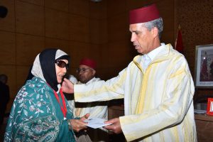 Man in a cream and yellow traditional robe (fez) presents a document to a woman in a teal floral shawl and hijab at a formal event, indoors with wood panel walls nearby.