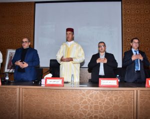 Four men stand behind a conference table during a formal event; the center man wears a yellow traditional Moroccan djellaba and fez while others in suits hold their hands to their chests.