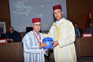 Two men in red fez and striped djellabas exchange a blue gift bag during a formal ceremony, with officials seated behind them.