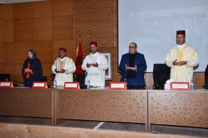 Formal panel at a wooden conference table; five people stand, some in traditional Moroccan robes, a Moroccan flag in the background.