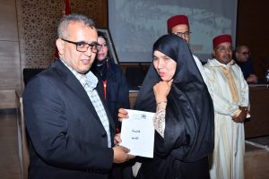 Man in a dark blazer and glasses hands a certificate to a woman in a black hijab during an indoor ceremony, with attendees in the background.