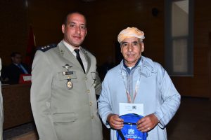 Military officer in a beige dress uniform with medals poses with an older man in a light blue traditional robe and turban, holding a blue backpack indoors at an event.