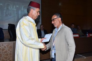 Man in a red fez and striped djellaba hands a certificate to a man in a beige jacket during a formal ceremony, shaking hands on stage.