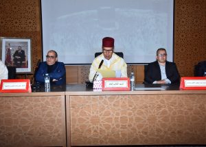 Panel of three men seated at a conference desk; center man in traditional Moroccan attire and fez reads from papers, with microphones and nameplates nearby.