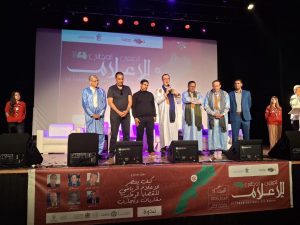 Group of men and women in traditional and formal attire standing on a stage during an event, with a large banner and speakers in the background.
