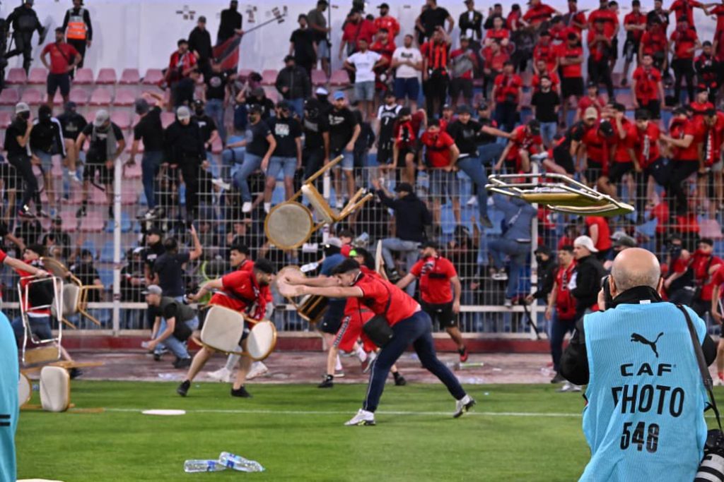 Crowd of fans on a football field throwing chairs with a photographer in a blue vest filming from the sideline.