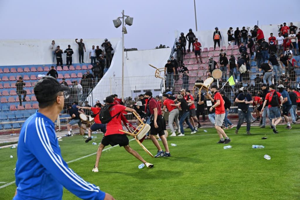Crowd of protesters on a stadium field throwing chairs while others run across the grass, stands filled with spectators in the background and scattered litter on the ground.