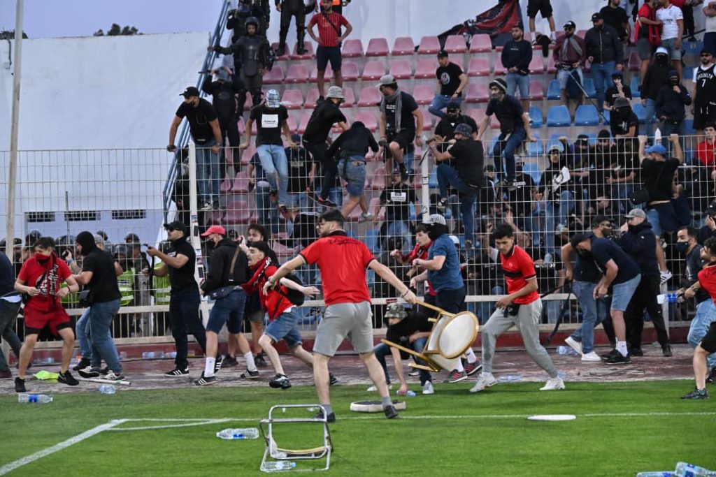Crowd of stadium spectators in red and black scattered around a metal barrier as people run and push during a chaotic brawl; a drum lies on the ground near the foreground