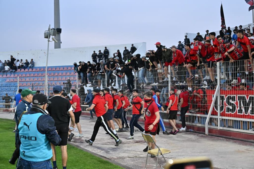 Soccer players in red shirts walk along the sideline as a crowd of red-clad fans cheers behind a fence on the stands.