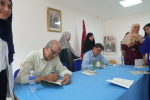 Two men sit at a blue-clothed table signing books as a group of women watch in a bright room with portraits on the wall, suggesting a book signing event.