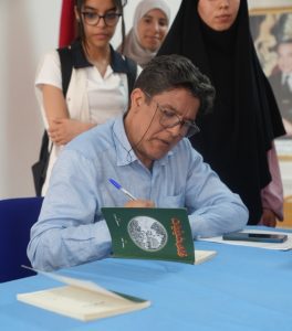 Man in a light blue shirt signs a book at a table, with three women in hijabs standing behind him.