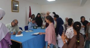 People line up at a table where two men sign documents during a community event in a bright room with a blue tablecloth.