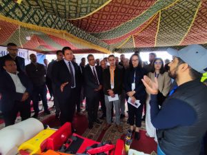 Group of professionals in business attire listening to a speaker under a colorful fabric tent, with safety tools on a table in the foreground.