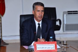 Man in a dark pinstripe suit seated at a conference table, speaking into a microphone, with a red nameplate in front (Arabic text).