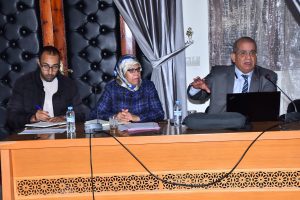 Three panelists at a conference table: left man writes notes, middle woman in a hijab listens, right man in a suit speaks with a laptop in front of him.