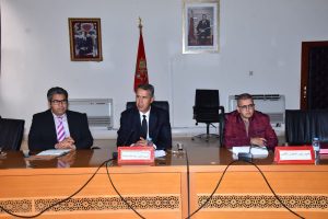 Three men in suits sit at a long conference table with microphones, papers, and water bottles during a formal meeting; a red flag stands behind them. Candid panel discussion setting in a government office.
