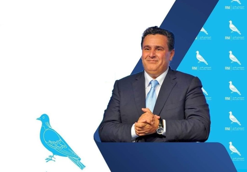 akhnouch-aziz | MCG24 | موقع إلكتروني مغربي شامل متجدد على مدار الساعة Man in a suit smiles while standing behind a blue podium at a press event with blue bird logos in the background.