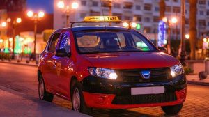 Red taxi car with roof sign driving through a lit city street at night, headlights on.