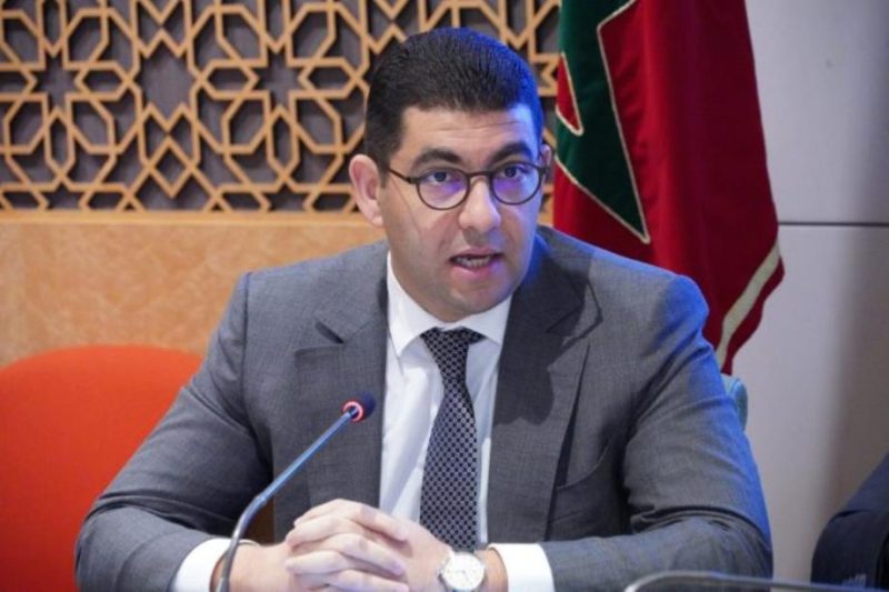 vis_0219202622410611 | MCG24 | موقع إلكتروني مغربي شامل متجدد على مدار الساعة Man in a gray suit and glasses sits at a conference table, speaking into a microphone with a patterned wall and flag in the background.
