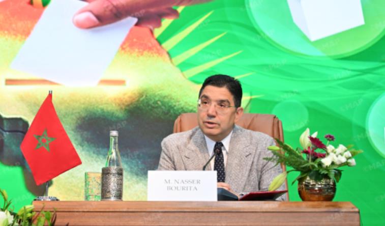 vis_0420202617333739 | MCG24 | موقع إلكتروني مغربي شامل متجدد على مدار الساعة Panelist at a wooden desk with Moroccan flag, nameplate reading M. Nassir Bourirat, a water bottle, and flowers on the table with a bright green backdrop.