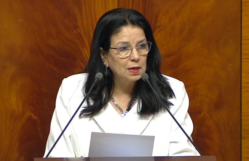 vis_0420202618283893 | MCG24 | موقع إلكتروني مغربي شامل متجدد على مدار الساعة Woman with black hair and glasses speaks into two microphones at a wooden podium, reading from a sheet.