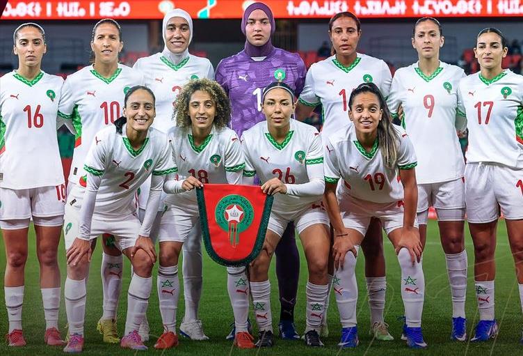 vis_0421202614502641 | MCG24 | موقع إلكتروني مغربي شامل متجدد على مدار الساعة Morocco women's national football team posing for a pre-match group photo, holding a green-and-red shield banner on the field.