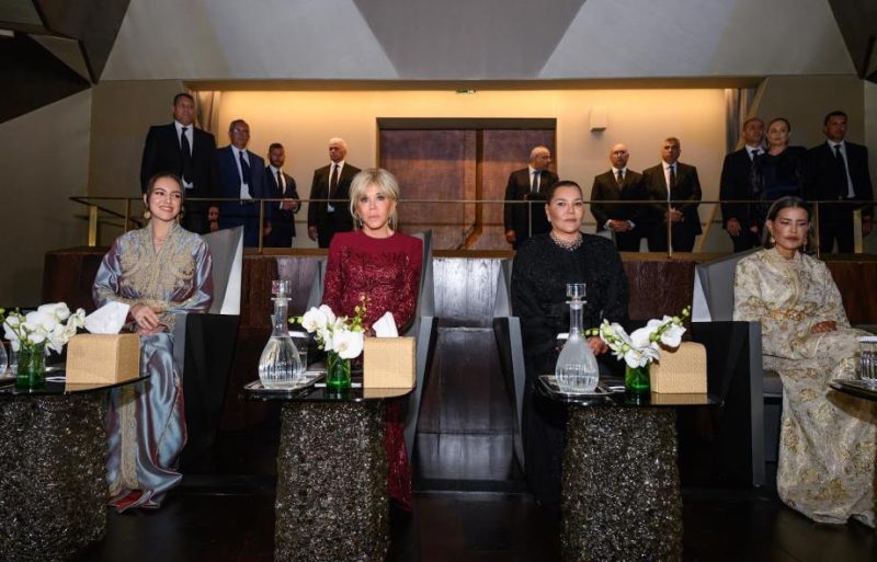 vis_0422202622180407 | MCG24 | موقع إلكتروني مغربي شامل متجدد على مدار الساعة Four women in elegant gowns sit at a formal event table with flowers; suited guests stand behind them.