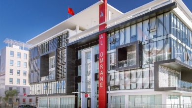 مجموعة-العمران | MCG24 | موقع إلكتروني مغربي شامل متجدد على مدار الساعة Modern glass-fronted building with a tall red column reading 'AL OMRANE' against a clear blue sky.