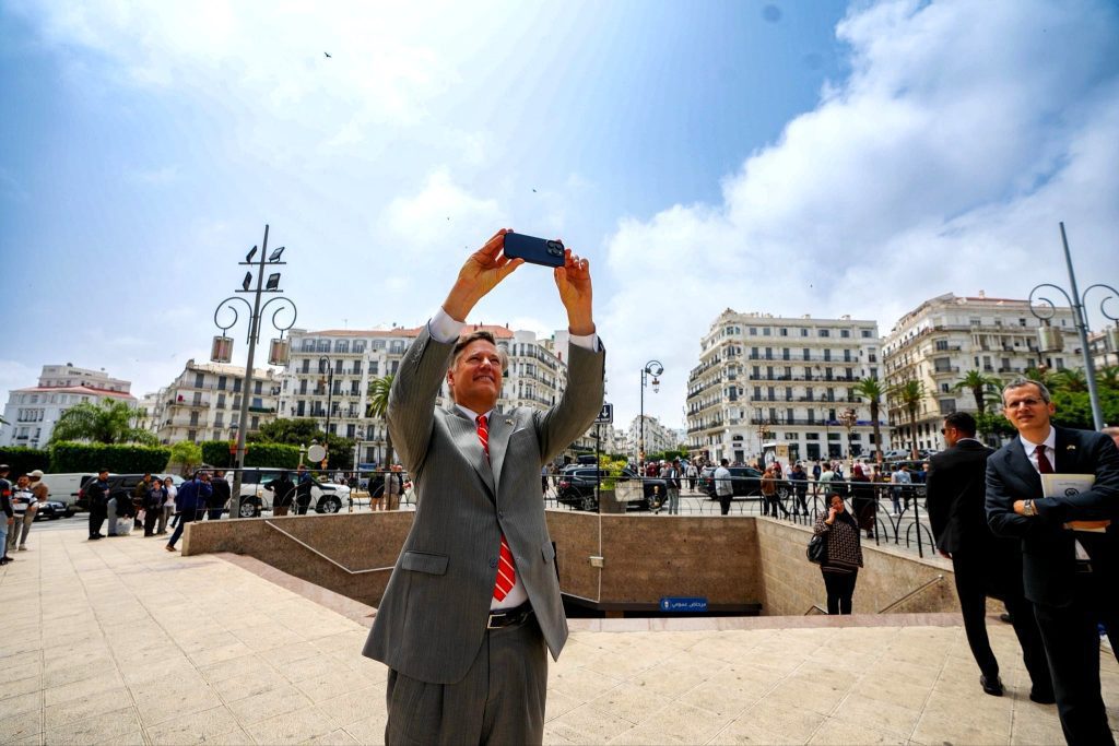 نائب وزير الخارجية الأمريكي | MCG24 | موقع إلكتروني مغربي شامل متجدد على مدار الساعة Man in a light gray suit and red tie holds a phone overhead to take a selfie in a busy city square with onlookers and white buildings behind him.