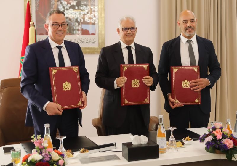 1778785871800 | MCG24 | موقع إلكتروني مغربي شامل متجدد على مدار الساعة Three men in dark suits stand at a conference table, each holding a red folder with a gold emblem during a formal ceremony.