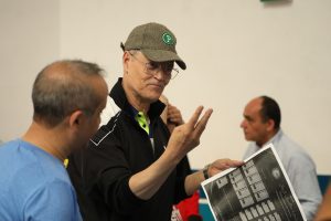 Man wearing a tweed cap and glasses explains a large printed chart to others in a group setting.