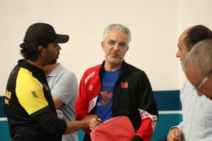 1P2A0025 | MCG24 | موقع إلكتروني مغربي شامل متجدد على مدار الساعة Middle-aged man with gray hair and glasses wearing a red and black jacket, talking with others in a casual group setting.