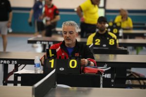 Male table tennis official seated at a score table showing 1–0, players blurred in the background