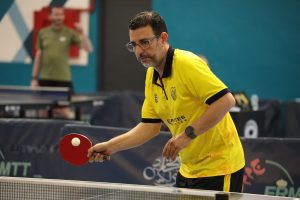 Table tennis player in a yellow shirt prepares to hit a white ball with a red paddle near the net indoors.