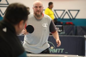 Bearded man in a gray shirt holding a table tennis paddle during a game in an indoor gym.